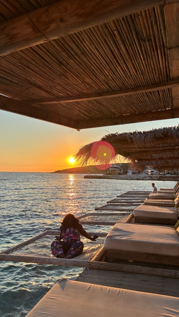 Une personne assise sur une plateforme au bord de l'eau, regardant le coucher de soleil sur la mer, avec des sièges en bois en arrière-plan.