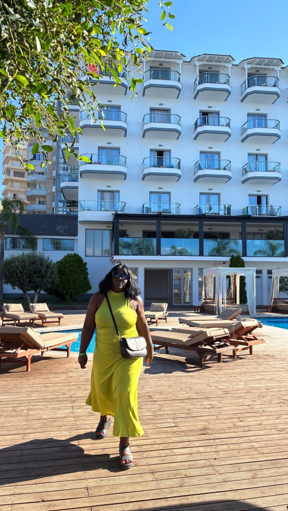 Une personne marchant sur un pont en bois près d'une piscine, devant un hôtel moderne avec plusieurs balcons, sous un ciel bleu clair.