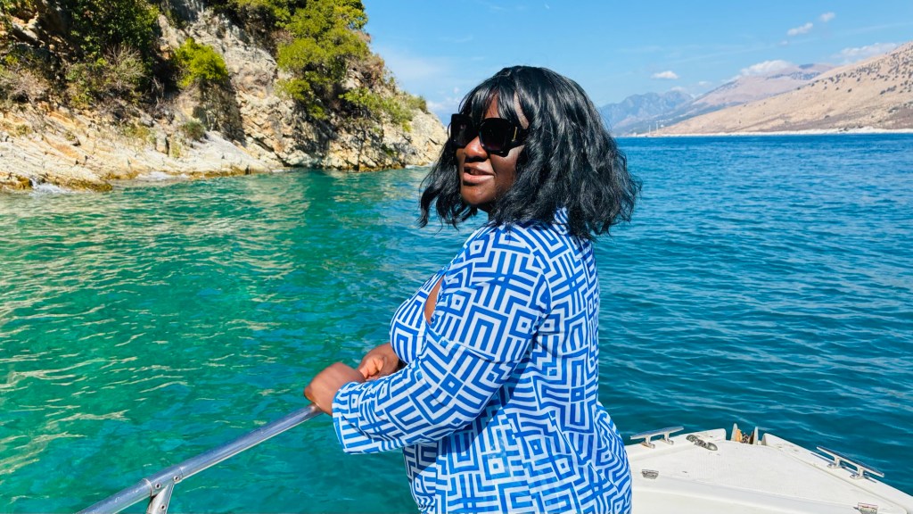Femme noire portant des lunettes de soleil, souriante, sur un bateau au bord d'une mer turquoise avec des montagnes en arrière-plan.