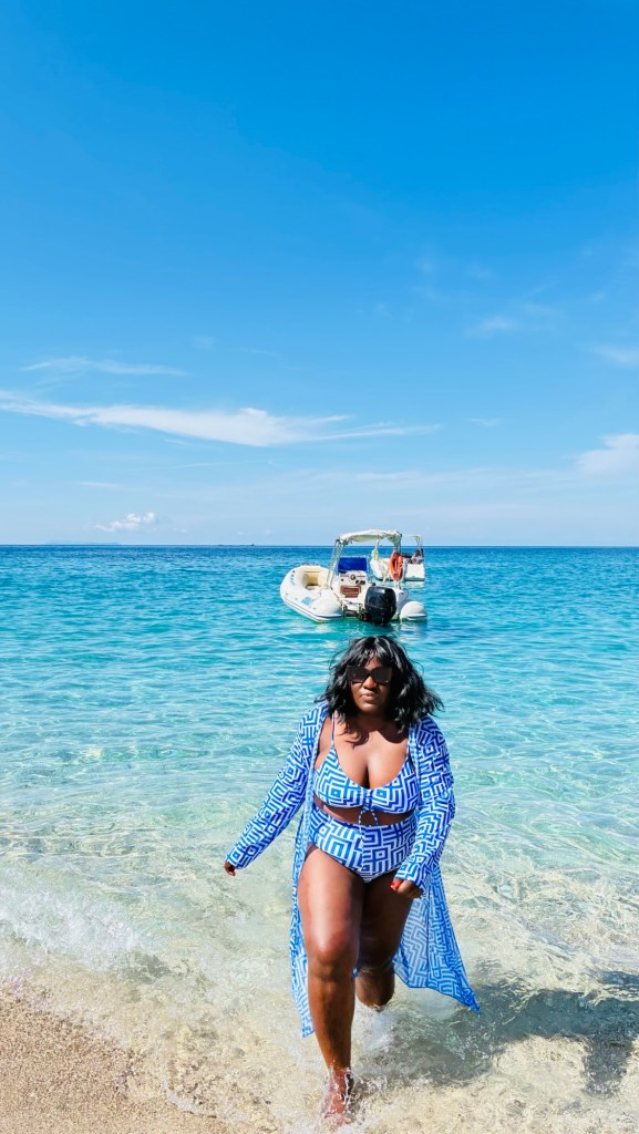 Une femme marche sur la plage, vêtue d'un maillot de bain et d'une couverture, avec un bateau à moteur au loin sous un ciel bleu clair.
