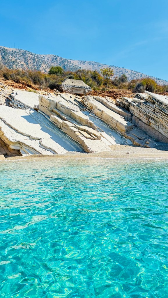 Vue d'une plage avec des roches blanches et des eaux turquoise claires, surplombée par des collines verdoyantes sous un ciel bleu.