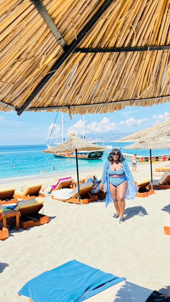 Une personne en maillot de bain marche sur une plage, sous un parasol en paille, avec des chaises longues autour et des bateaux au loin.
