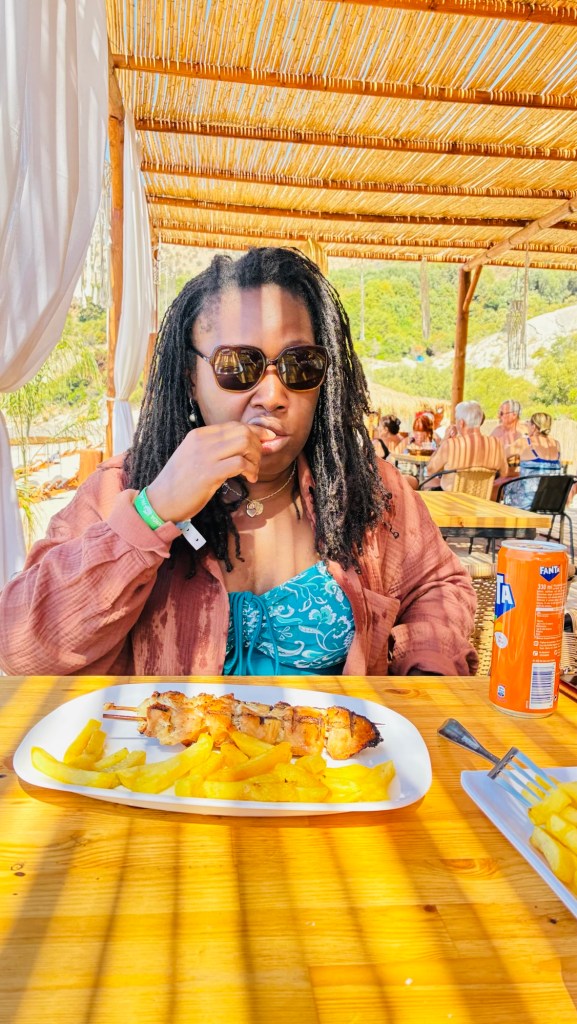 Une personne assise à une table en bois, portant des lunettes de soleil et un haut bleu, dégustant un plat de brochettes grillées avec des frites. En arrière-plan, plusieurs personnes profitent d'un cadre en plein air.