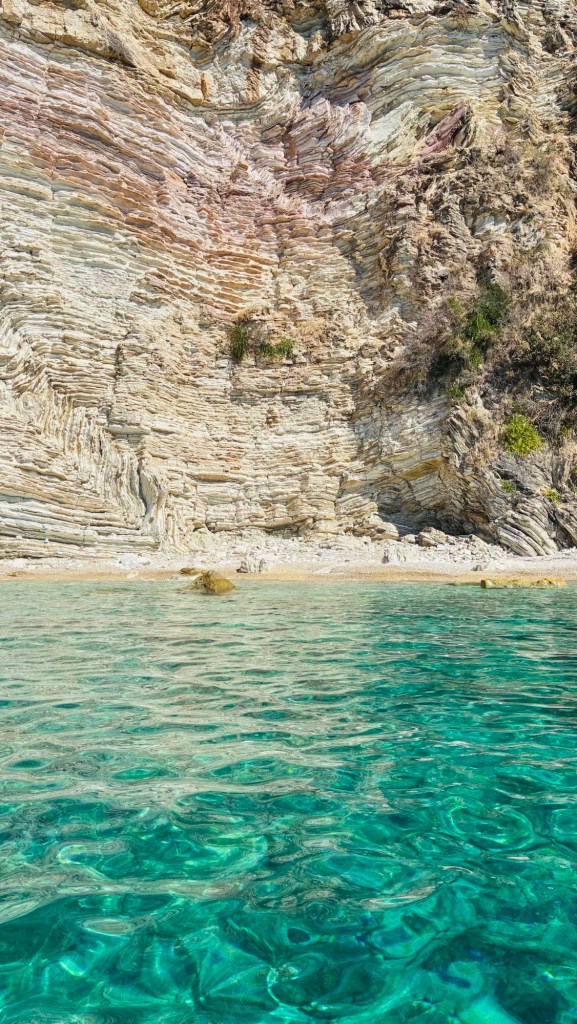 Vue d'une eau cristalline près d'une falaise aux strates multicolores, avec du sable visible en bas.