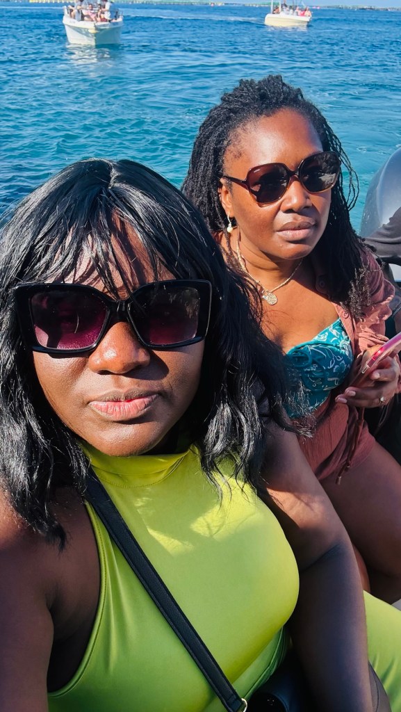 Deux femmes souriantes posent pour un selfie en bateau, avec des lunettes de soleil, devant une mer bleue et des bateaux en arrière-plan.