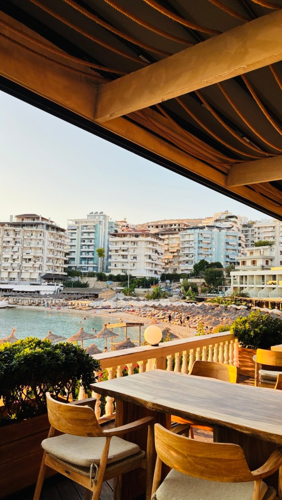 Vue sur une plage depuis une terrasse, avec des chaises en bois et une table en bois, entourée d'arbustes et d'immeubles en arrière-plan.