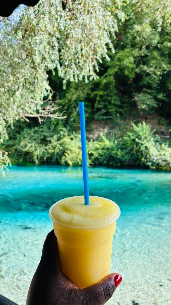 Un verre de smoothie jaune avec une paille bleue, tenu dans une main, devant un paysage naturel avec de l'eau claire et des arbres en arrière-plan.