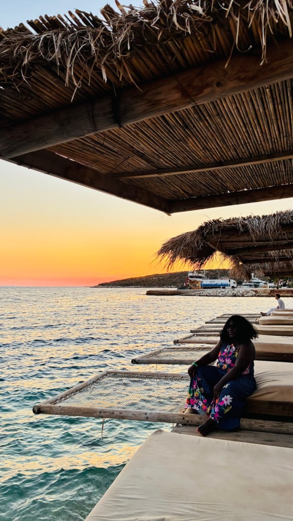 Une femme assise sur une plateforme au bord de l'eau, entourée de parasols en paille, avec un coucher de soleil vibrant en arrière-plan.