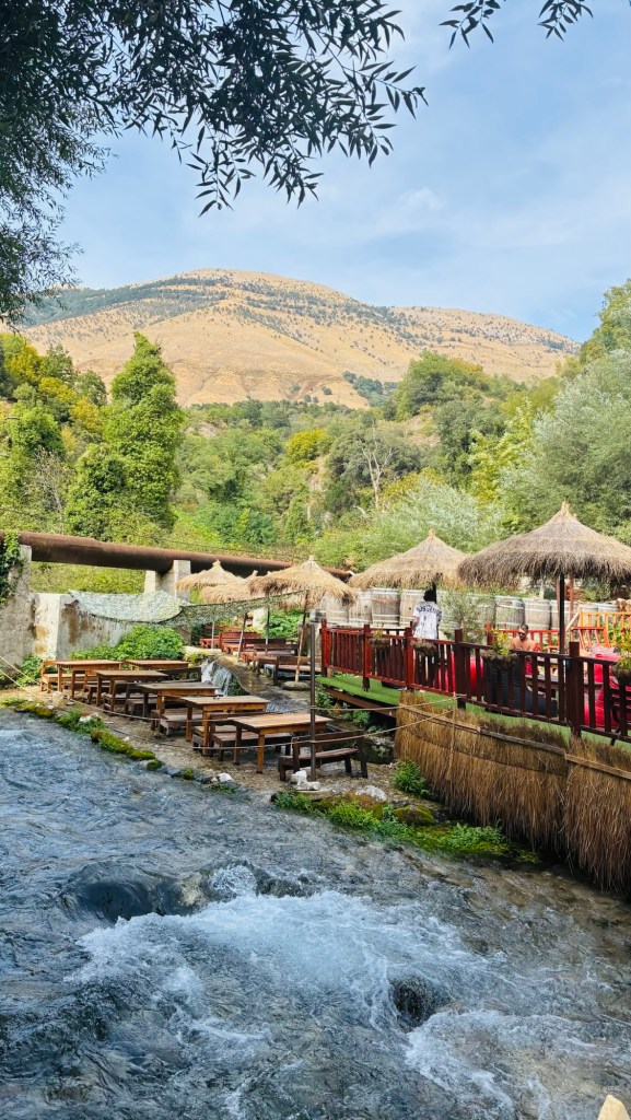 Un ruisseau clair coule à travers un paysage verdoyant, avec des tables en bois et des parasols en chaume visibles sur une terrasse près de l'eau. En arrière-plan, des collines d'un jaune doré se dressent sous un ciel bleu avec quelques nuages.