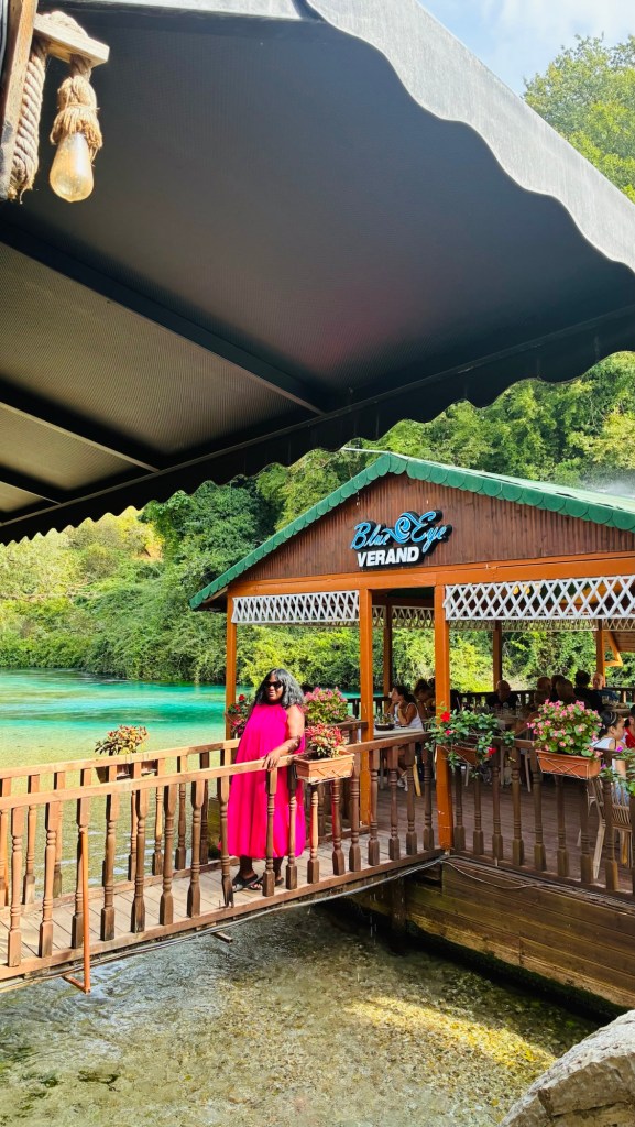 Une femme en robe rose se tient sur un pont en bois menant à un restaurant nommé 'Blue Eye Verand', entouré de verdure et d'eau turquoise.