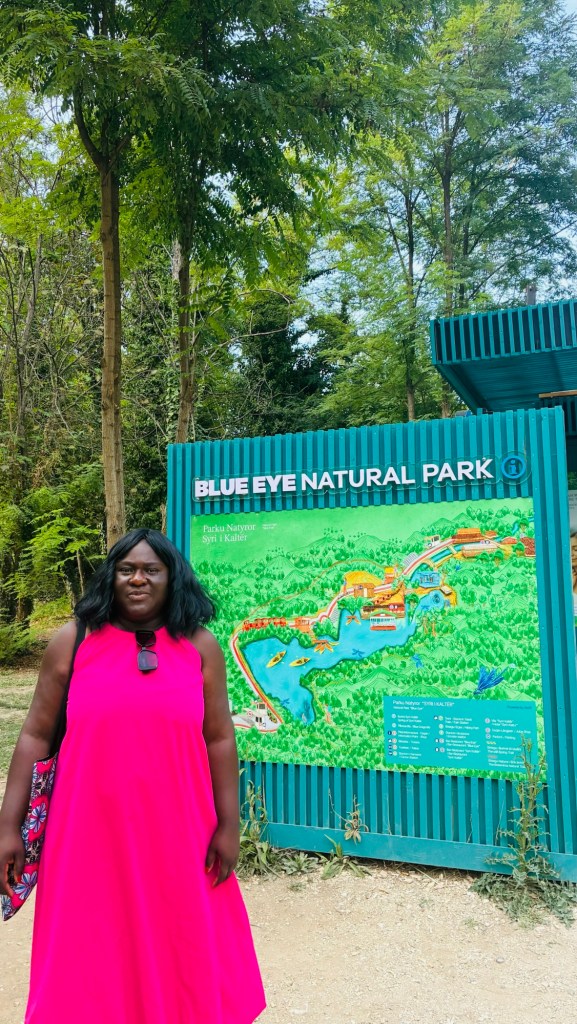 Femme souriante en robe rose se tenant devant un panneau du parc naturel Blue Eye, avec une carte détaillant les attractions et les sentiers du parc, entourée d'arbres.