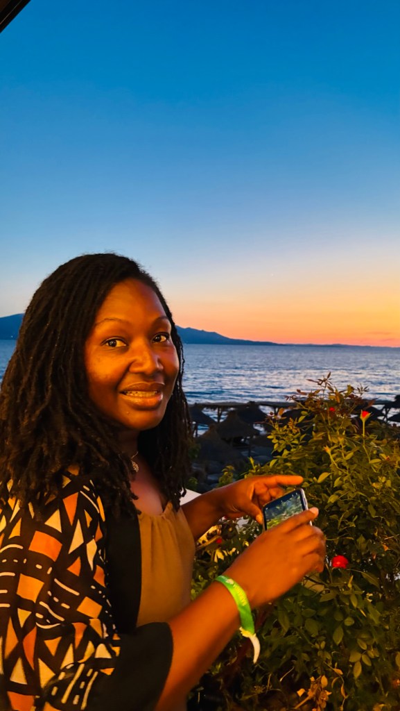 Une femme souriante avec des cheveux bouclés se tient devant un coucher de soleil au bord de la mer, tenant un téléphone et entourée de fleurs.