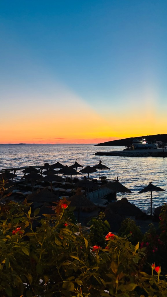 Coucher de soleil sur la mer, avec des parasols en bord de plage et des fleurs colorées au premier plan.