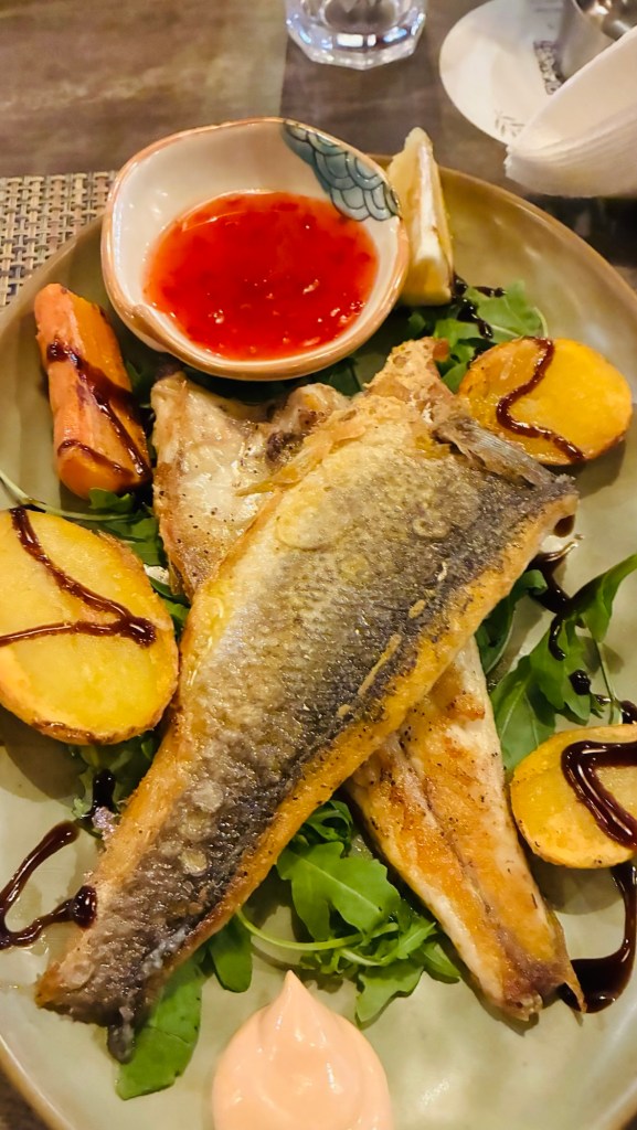 Un plat de poisson grillé servi avec des morceaux de patates, des carottes, une sauce aigre-douce et un quartier de citron sur un lit de roquette.