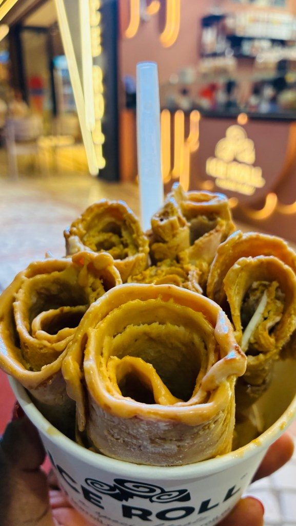 Bol de rouleaux de glace avec garniture crémeuse, tenu à la main. En arrière-plan, un comptoir de café avec des lumières décoratives.