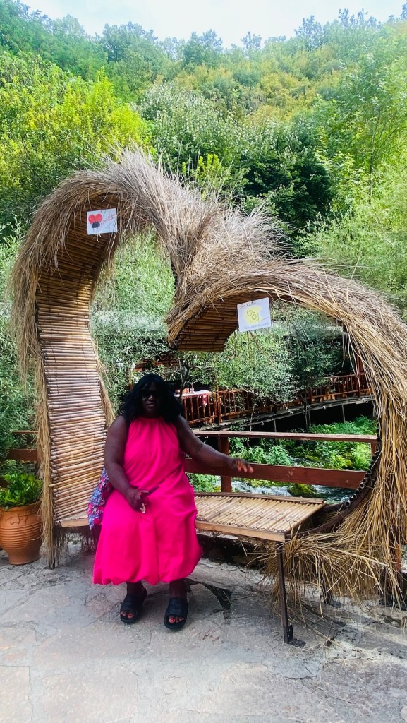 Femme assise sur un banc en bois sous une arche en paille dans un environnement verdoyant. Elle porte une robe rose et a des cheveux bouclés.