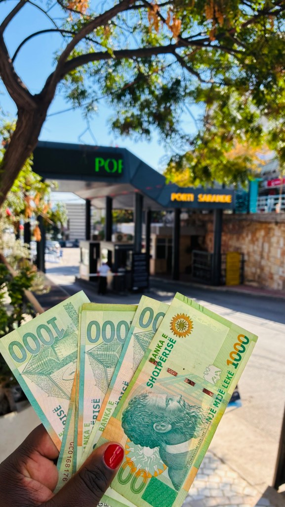 Une main tenant des billets de banque de 1000 unités devant une entrée de magasin portant le nom 'Porti Sarake'.