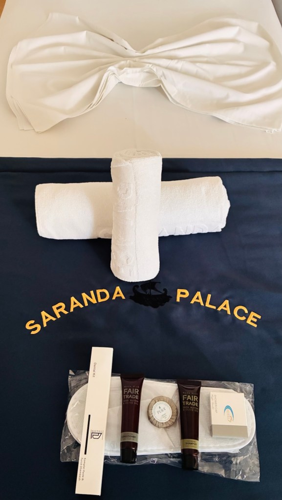 Vue d'un lit avec des serviettes blanches enroulées et des produits de soins sur une couverture à l'effigie de Saranda Palace.