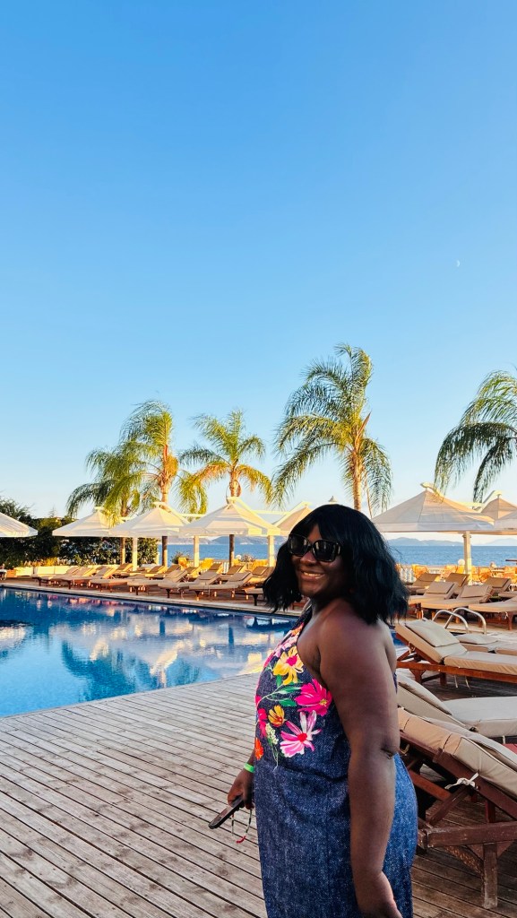 Une femme souriante, portant une robe à fleurs, se tient près d'une piscine entourée de chaises longues et de palmiers, sous un ciel bleu clair.