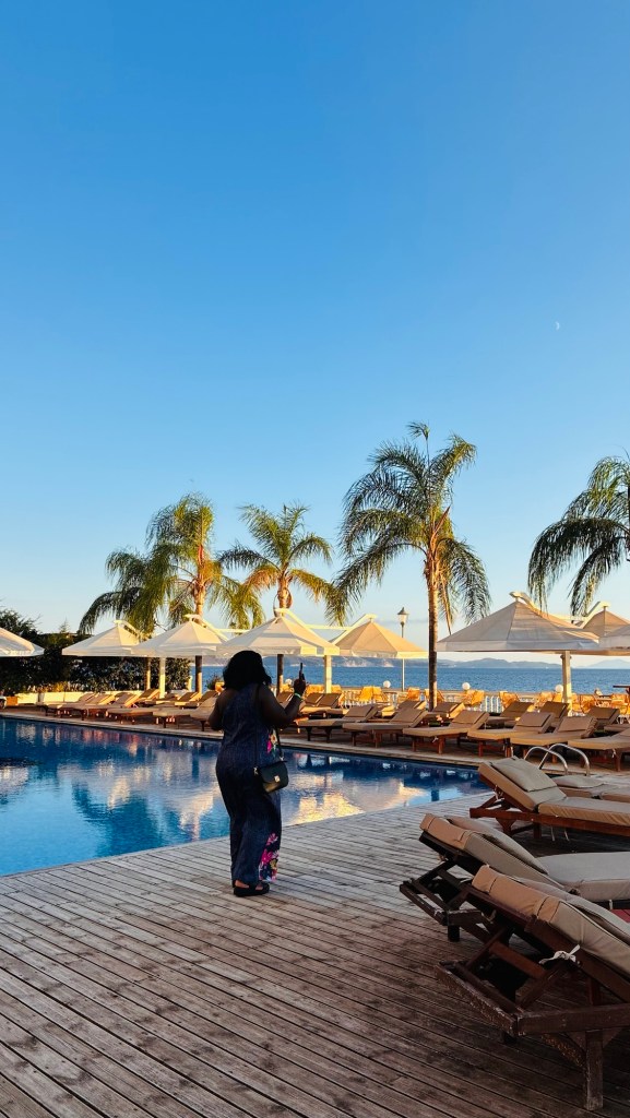 Une personne marchant au bord d'une piscine, entourée de transats et de palmiers, sous un ciel bleu clair.