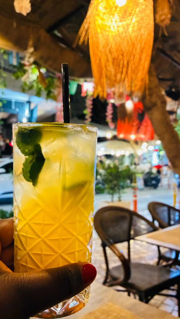 Un verre de cocktail jaune avec de la menthe, tenu par une main, en arrière-plan lumineux d'une terrasse de café.