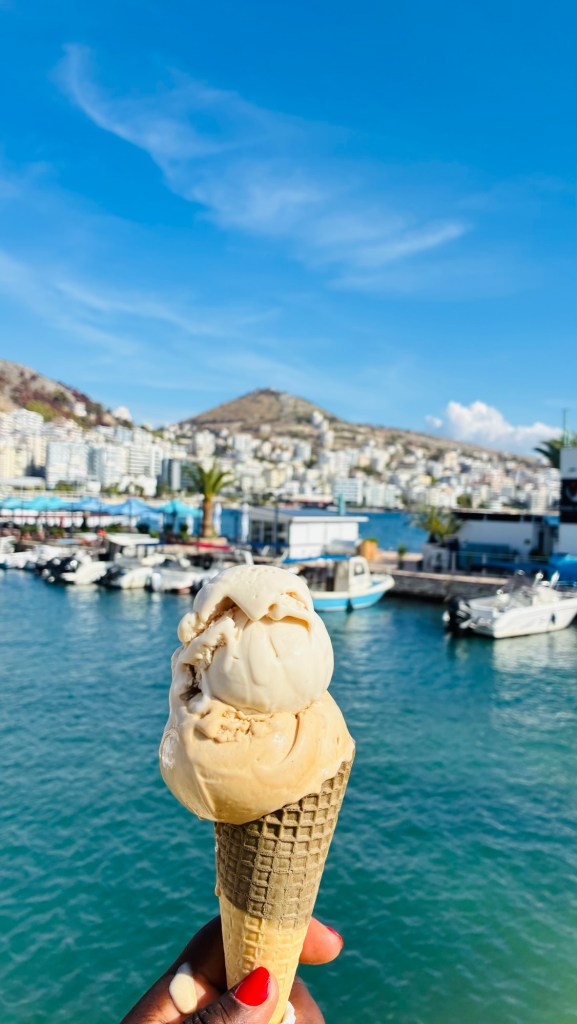 Cone de glace avec deux boules de crème glacée à l'avant-plan, avec un paysage de mer et une ville en arrière-plan.