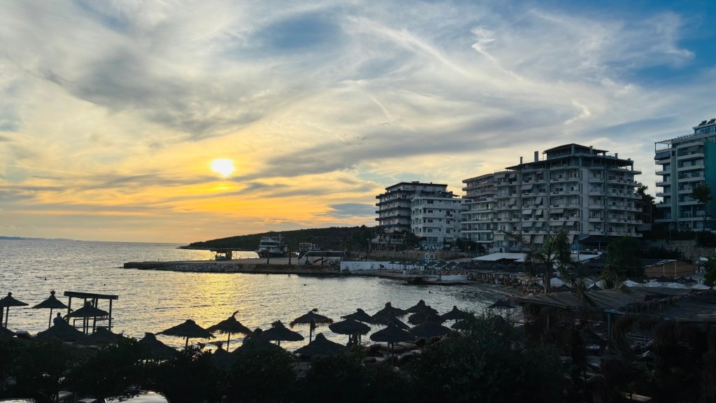 Coucher de soleil sur un bord de mer, avec des bâtiments modernes et des parasols en dessous.