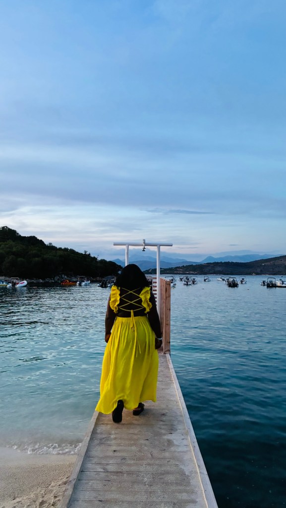 Une personne en robe jaune marche sur un quai en bois, face à l'eau, avec des bateaux au loin sous un ciel bleu clair.