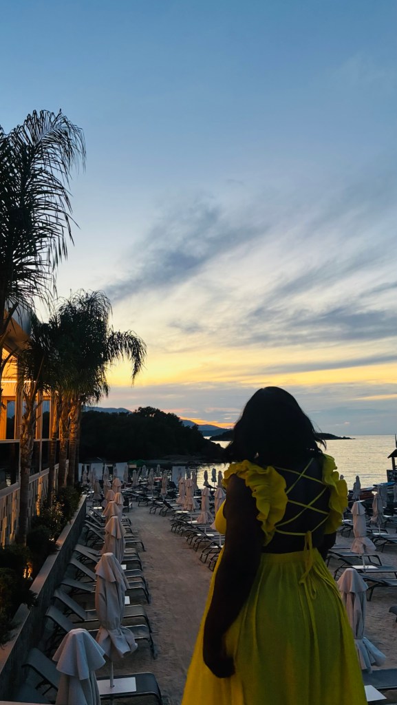 Une femme portant une robe jaune se tient sur une plage au coucher du soleil, entourée de transats et de parasols.