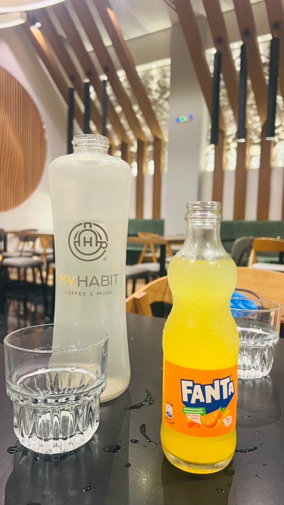 Une bouteille d'eau marquée MYHABIT, une bouteille de Fanta orange et deux verres d'eau sur une table dans un café moderne.