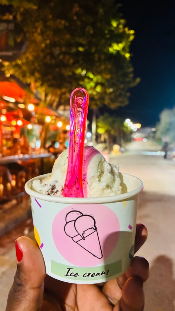 Un cornet de glace avec une cuillère en plastique rose, tenu par une main, de nuit, avec un décor urbain en arrière-plan.