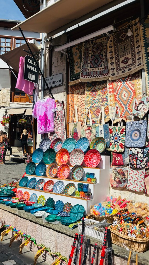 Un marché animé avec des assiettes colorées, des sacs brodés et des vêtements suspendus. En arrière-plan, des tapis décoratifs et des marchands attirent l'attention des visiteurs.