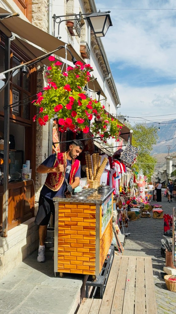 Un homme en costume traditionnel sert des glaces depuis un chariot en bois dans une rue pavée, bordée de boutiques et de fleurs suspendues.