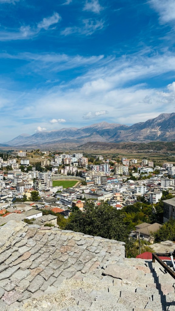 Vue panoramique d'une ville entourée de montagnes, avec des bâtiments et un terrain de sport visible en bas, sous un ciel bleu parsemé de nuages.