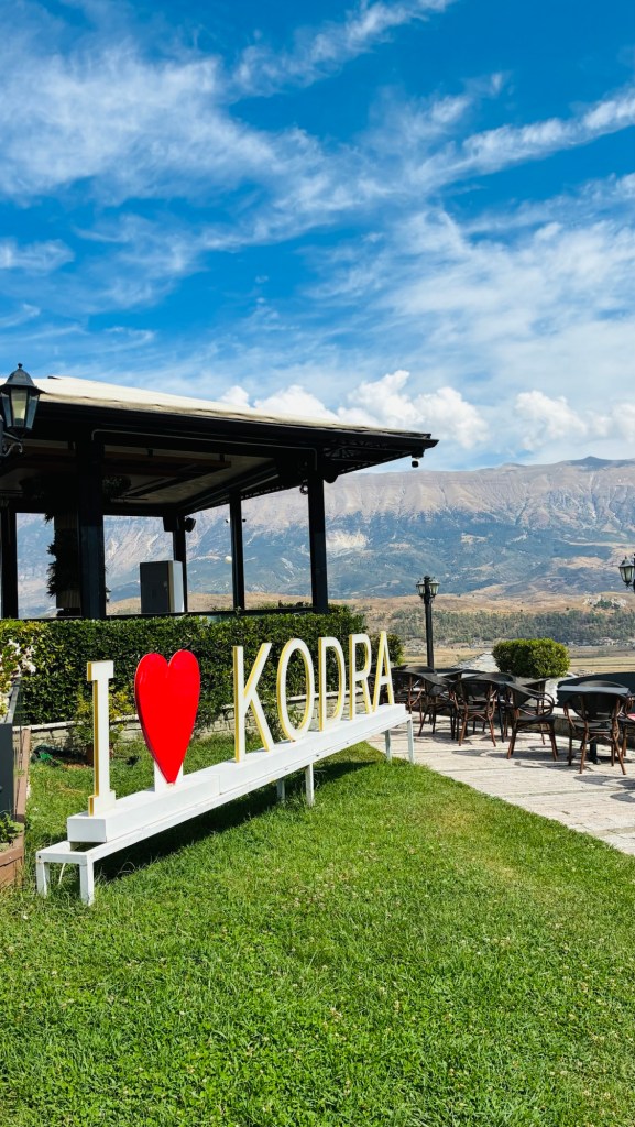 Panneau 'I ❤️ KODRA' en lettres blanches sur une pelouse verte, avec un ciel bleu et des montagnes en arrière-plan.