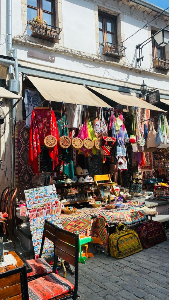 Un marché coloré avec des étals remplis d'artisanat, de vêtements traditionnels et de décorations suspendues, devant un bâtiment à deux étages avec des fenêtres à balcons.