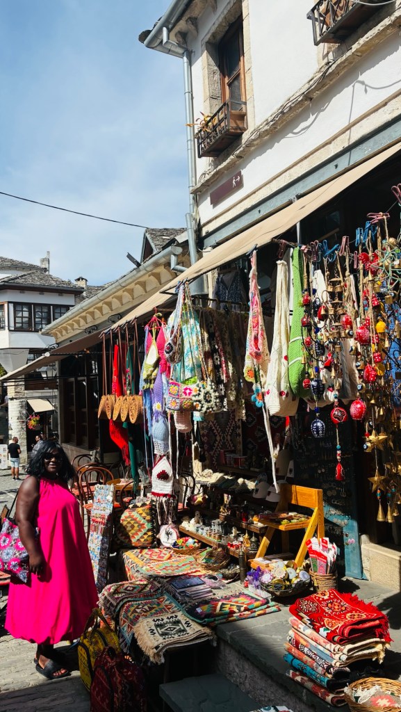 Femme portant une robe rose se tenant devant un marché coloré avec des tapis et divers artisanats suspendus.