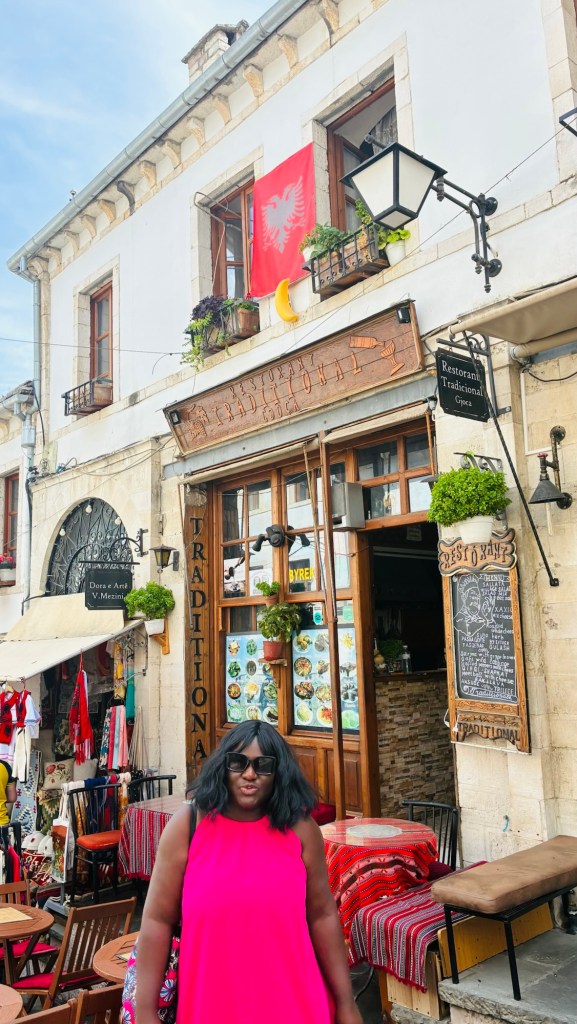 Une femme en robe rose se tient devant un restaurant traditionnel avec une enseigne en bois, des drapeaux et des plantes en pot, dans une rue animée.