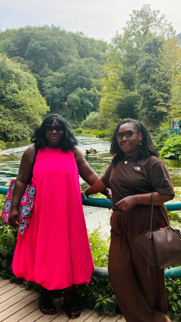 Deux femmes souriantes posent près d'une rivière entourée de verdure. L'une porte une robe longue rose, l'autre est habillée en marron. Elles portent des lunettes de soleil.